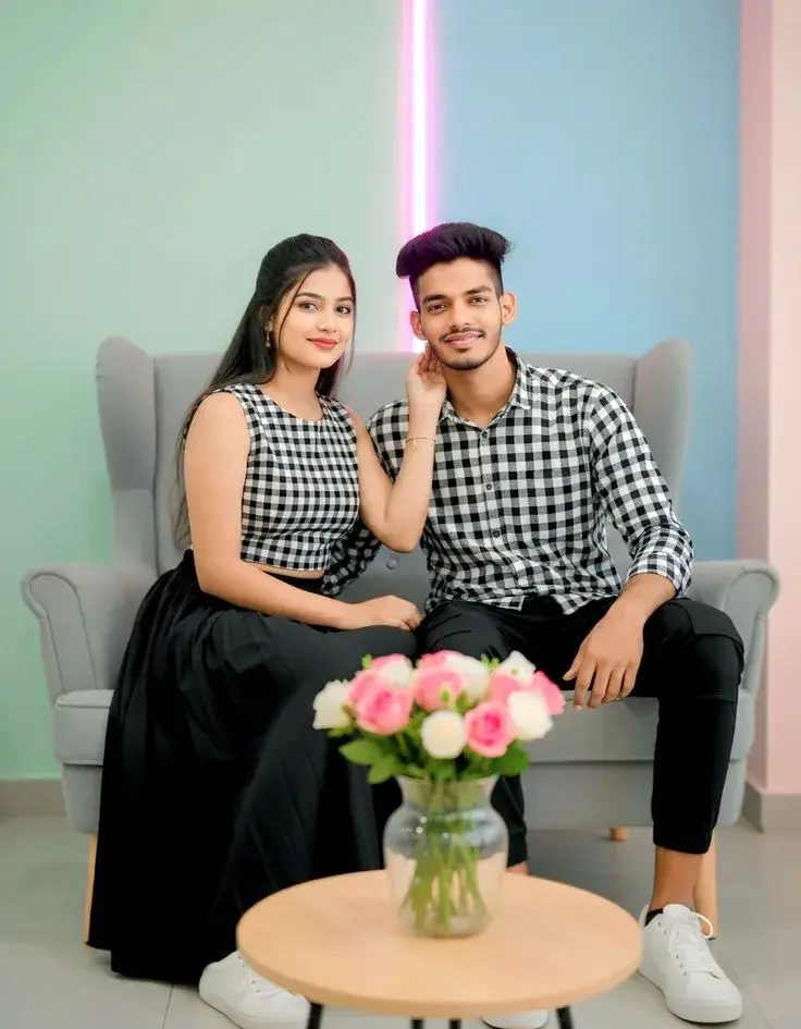 Candid Sunlit Studio Portrait Of Smiling Young South Asian Couple In Matching Checkered Outfits Seated On Grey Armchair