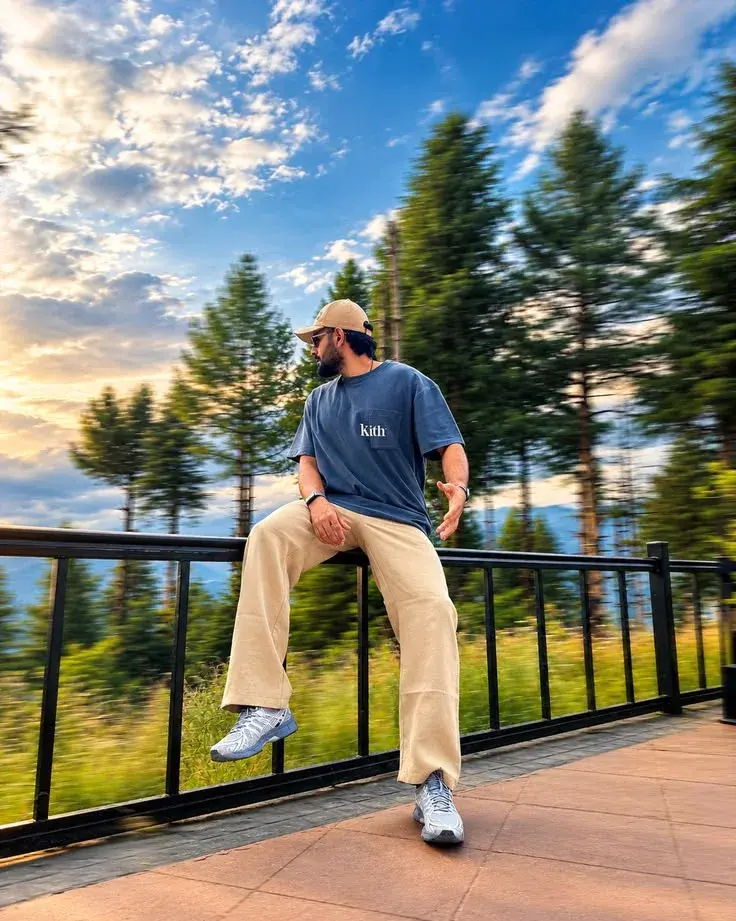 Candid Sunset Terrace Portrait Of South Asian Man In Kith T-Shirt And Beige Wide Trousers Perched On Balcony Railing