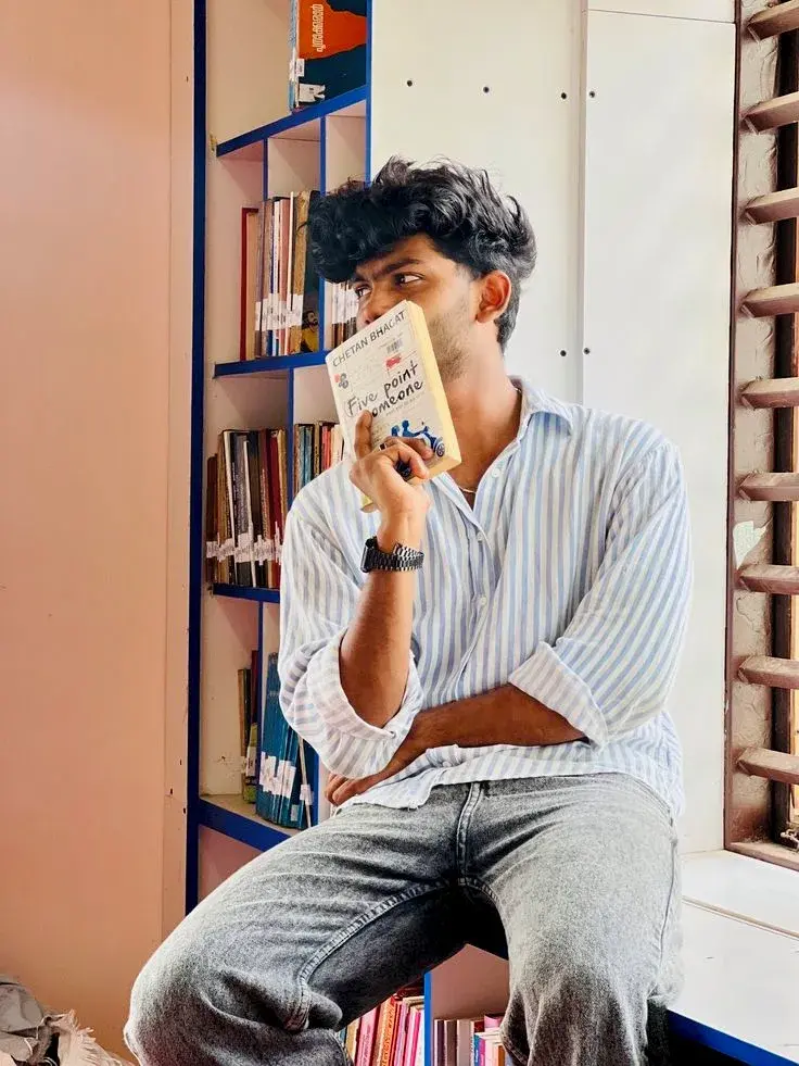 Candid Window Seat Portrait Of Young South Asian Man Holding Paperback Book In Library Corner