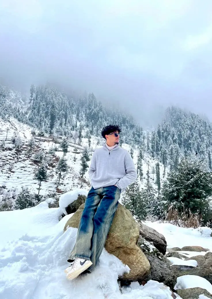 Candid Winter Mountain Portrait Of Young South Asian Man Leaning On Bolder In Snow-Covered Alpine Forest