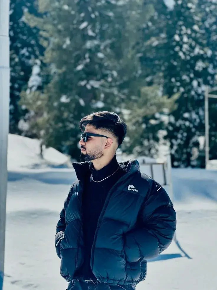 Candid Winter Portrait Of Young South Asian Man In Black Puffer Jacket And Sunglasses Against Snowy Pine Mountains