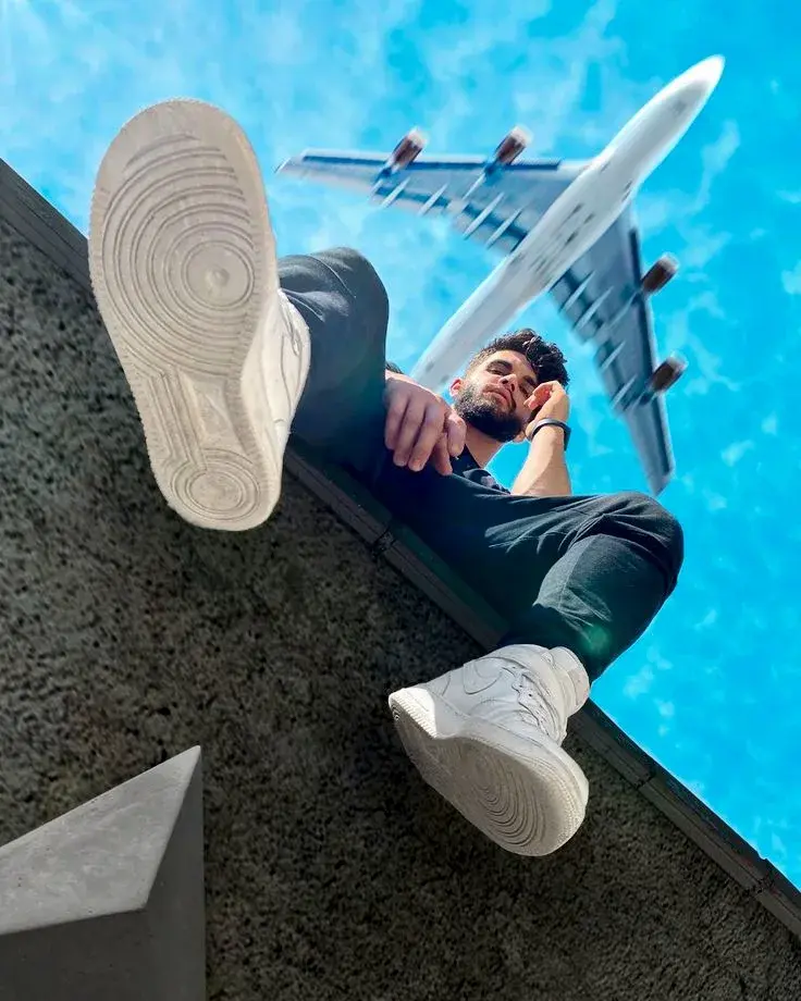 Dramatic Low-Angle Rooftop Portrait Of South Asian Man In Black Outfit And White High-Top Sneakers With Airplane Flying