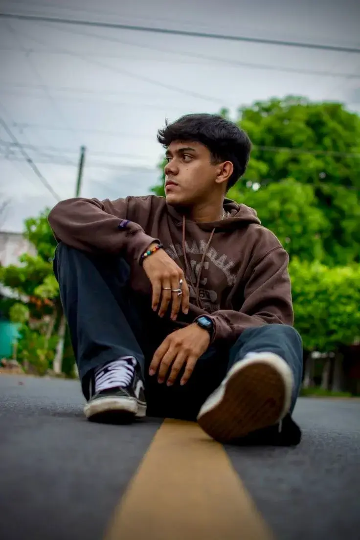 Low-Angle Streetwear Portrait Of Young South Asian Man Sitting On Asphalt Road With Yellow Center Line In Brown Hoodie A