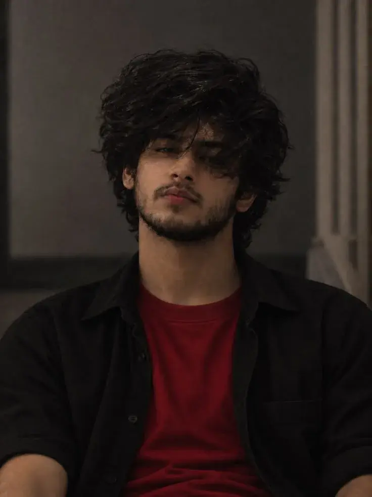 Moody Indoor Portrait Of Pensive Young South Asian Man With Shaggy Curly Black Hair In Red T-Shirt And Black Overshirt