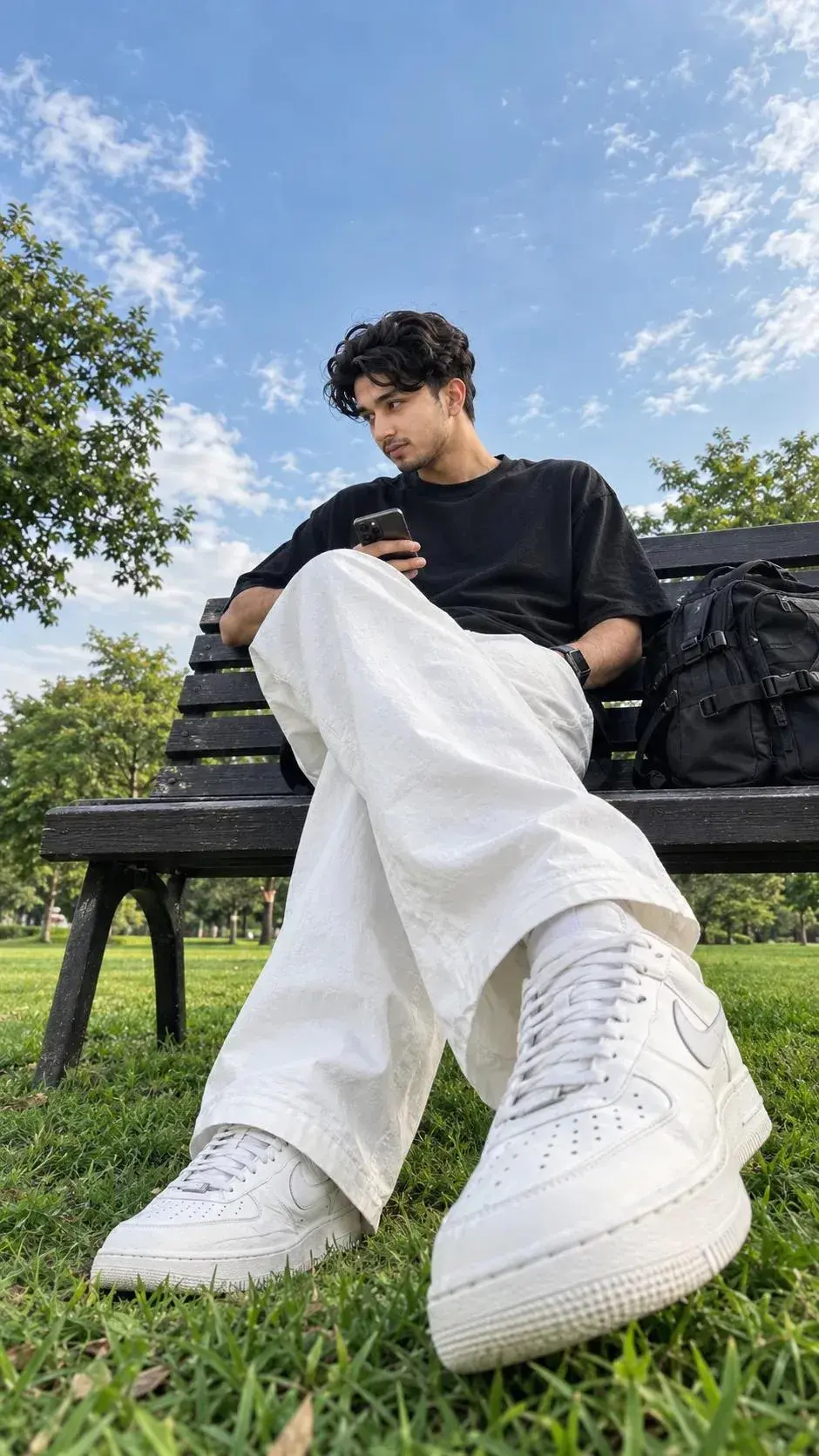Young Man In Black T-Shirt And White Baggy Pants Sitting On A Park Bench With Smartphone